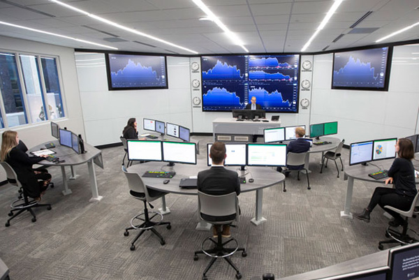 A modern classroom or trading‑lab setting where several people sit at curved desks working on multiple computer monitors, while an instructor stands at the front near large wall‑mounted screens displaying financial charts and data.