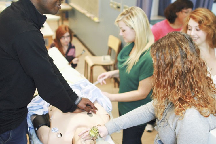 Group of people practicing a medical procedure on a training mannequin, using equipment such as sensors and a resuscitation bag in a classroom setting