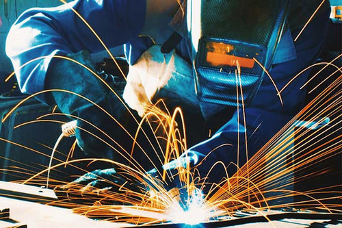 Person welding metal with bright sparks flying in multiple directions, wearing protective gloves and a dark welding helmet in an industrial workshop setting.