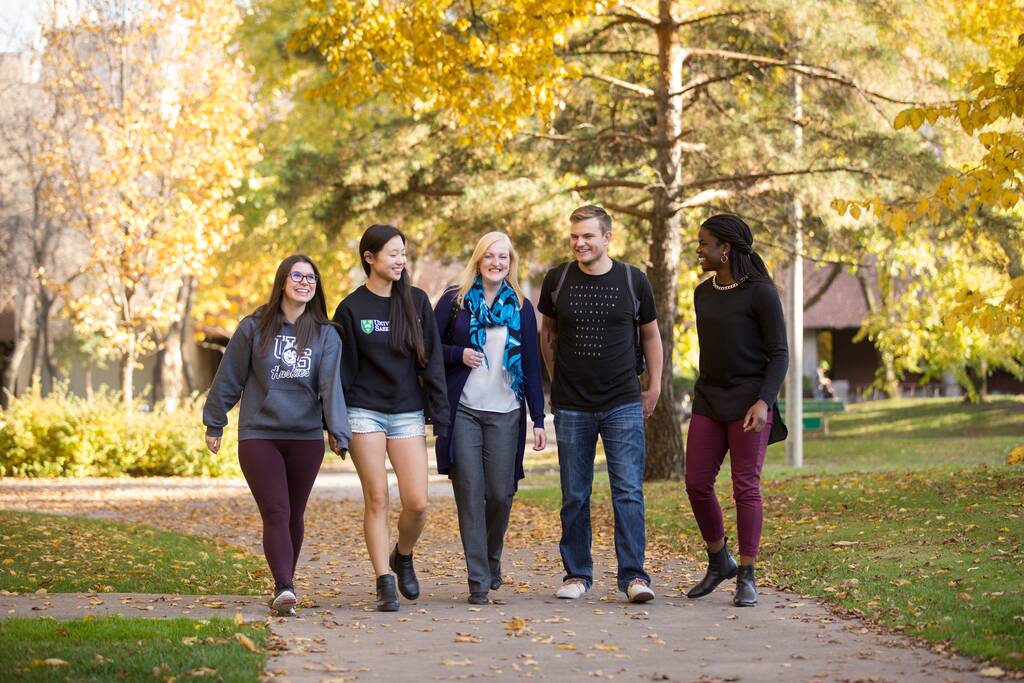 Five USask students chatting and laughing. It's fall. They walk down one of the pathways on the USask Saskatoon campus.