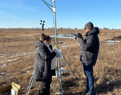 Two people install a weather monitoring mast in an open prairie research field.
