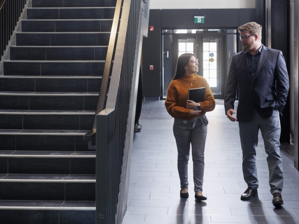 Two people walk through a building corridor beside a staircase, talking and holding a notebook.