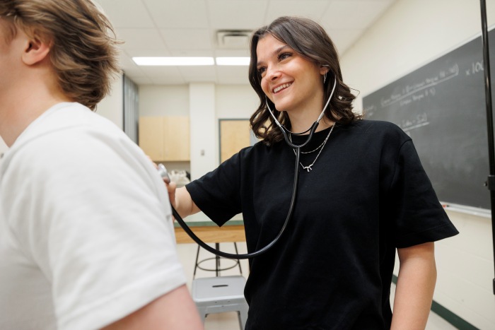 Students work at a lab table with anatomy models and laptops in a kinesiology classroom.