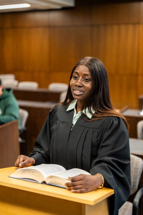 Student at podium reading from book in wood‑panelled law classroom.