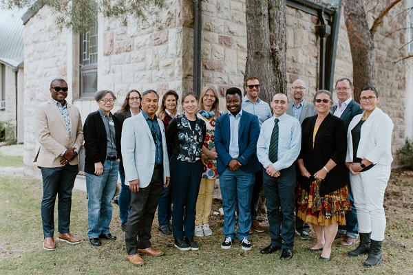 Group of adults standing together outdoors beside a stone building on a grassy area.