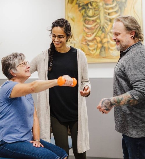 Seated patient lifts orange dumbbell as two clinicians coach in a clinic room