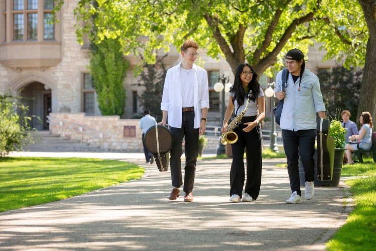Music students walk along a campus pathway carrying instrument cases and a saxophone.