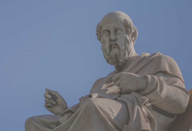 Stone statue of a seated philosopher holding an object against a clear blue sky.