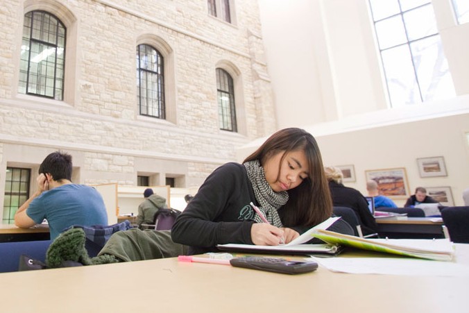 student writing at a table in a bright university library with other students studying nearby