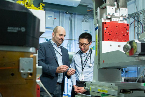 Two people in a laboratory examine a small component while standing beside large industrial research equipment with control panels, cables, and mechanical fixtures.