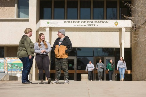 Three students talk and walk on a campus walkway in front of the College of Education building, while other students enter and exit the building in the background on a sunny day.