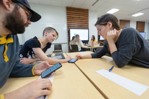 Students seated at tables in a classroom collaborate using smartphones and laptops, with a large screen displaying content in the background.