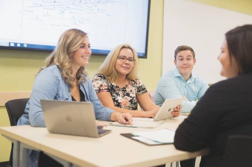 Group of people seated at a table in a classroom setting, using laptops and a tablet, with a large screen displaying a diagram in the background.