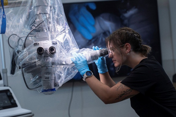 A healthcare trainee wearing gloves operates a medical imaging or surgical device covered in a protective plastic drape inside a clinical training lab, with monitors and medical equipment visible in the background.